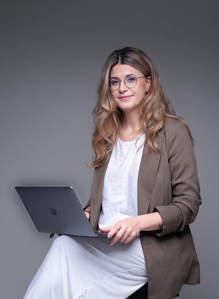 Portrett av en statsautorisert regnskapsfører med bærbar PC, smilende kvinne med briller i brun blazer på grå bakgrunn - Polkon Regnskap AS.